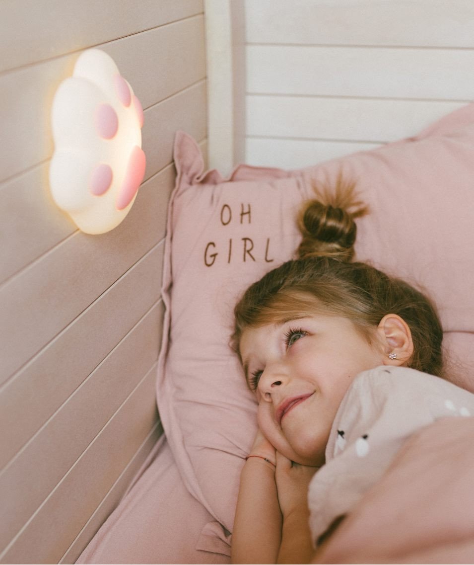 1. Young girl lying in bed smiling at wall-mounted pink paw-shaped silicone lamp in cozy children's room