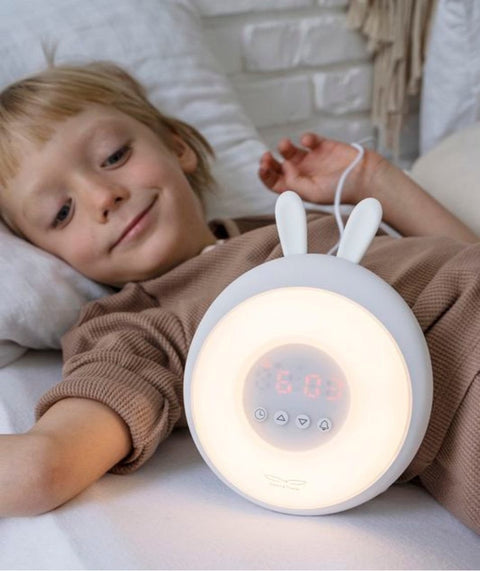 1. Child lying in bed with white Rabbit & Friends alarm clock lamp, showing warm light and rabbit ears