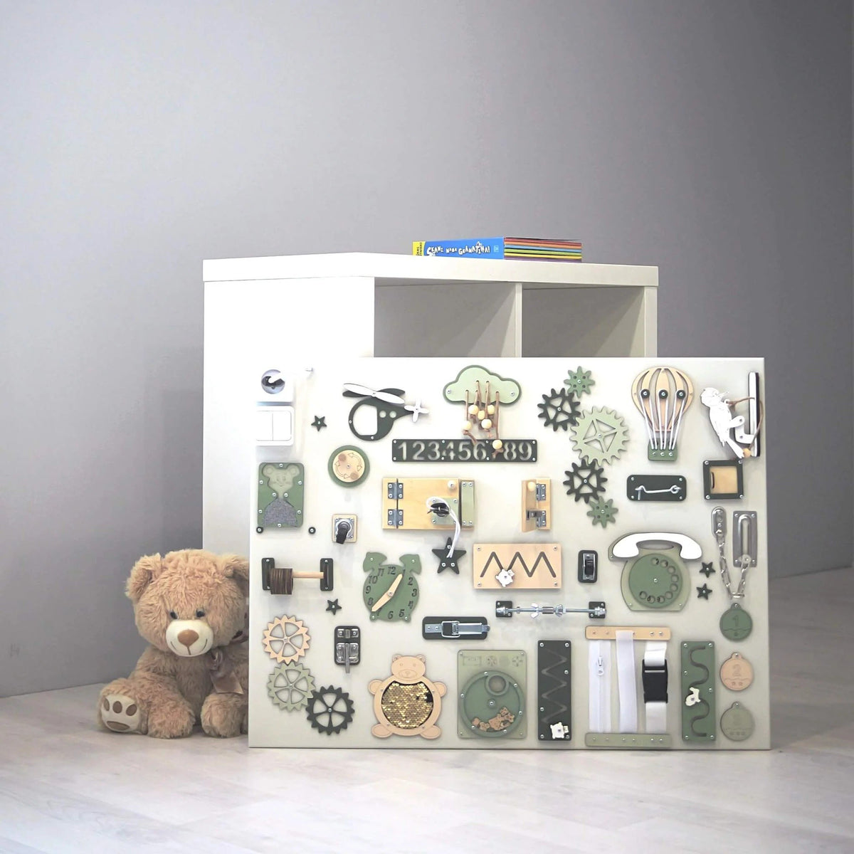 5. Montessori sensory busy board in green and beige, placed on the floor next to a teddy bear and a white shelf, ideal for children's playrooms.