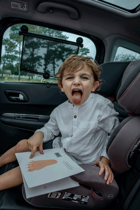 1. Child in car seat with Ezimoov sunshade on window, holding book and sticking out tongue
