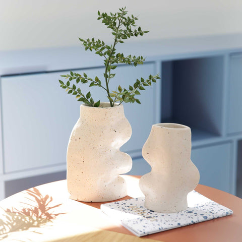 1. Lifestyle image of two Fluxo Ceramic Vases by 5mm Paper in white, showcasing unique flowing lines, placed on a table with greenery
