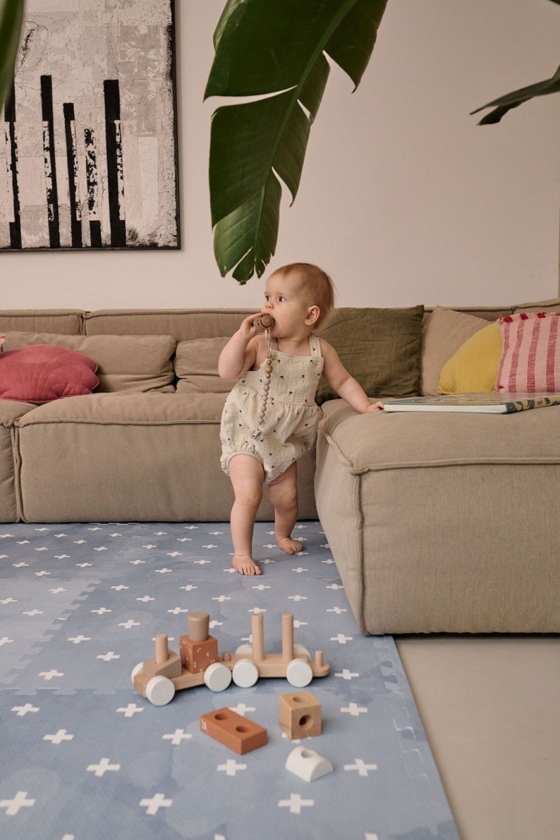 1. Baby standing by sofa on OLYA KIDS Cross Azure foam puzzle playmat with wooden toys