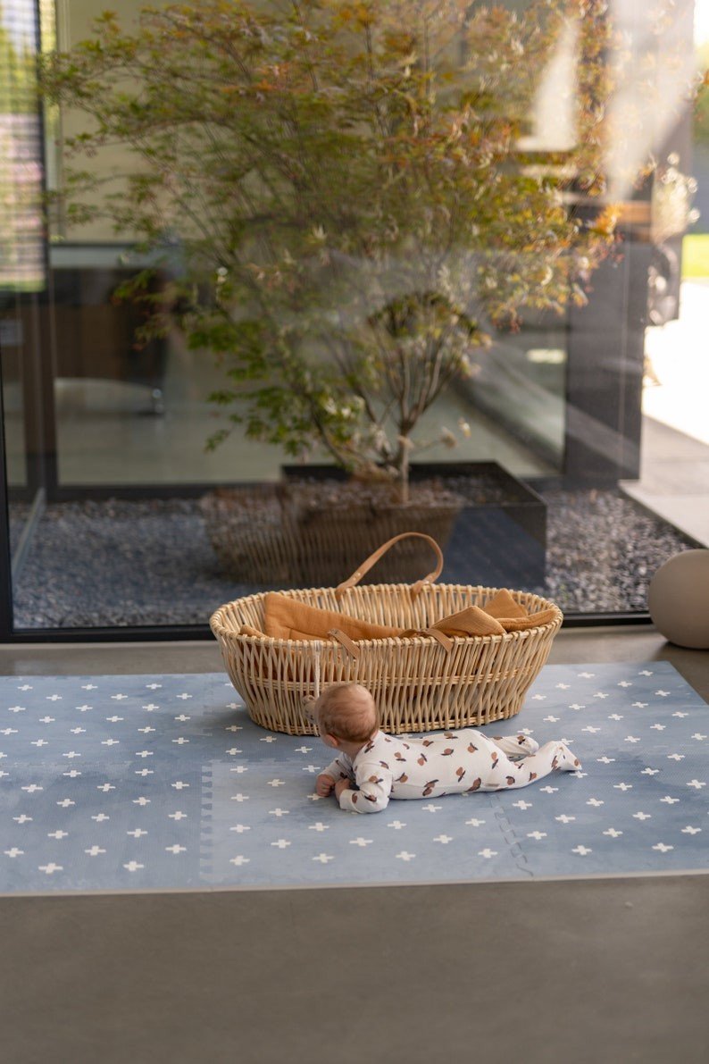 1. Baby lying on OLYA KIDS Cross Azure foam puzzle playmat next to wicker basket in sunlit room