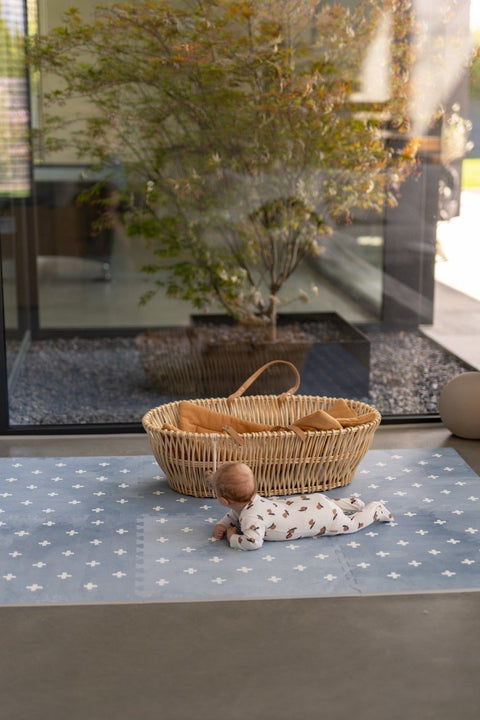 1. Baby lying on OLYA KIDS Cross Azure foam puzzle playmat next to wicker basket in sunlit room