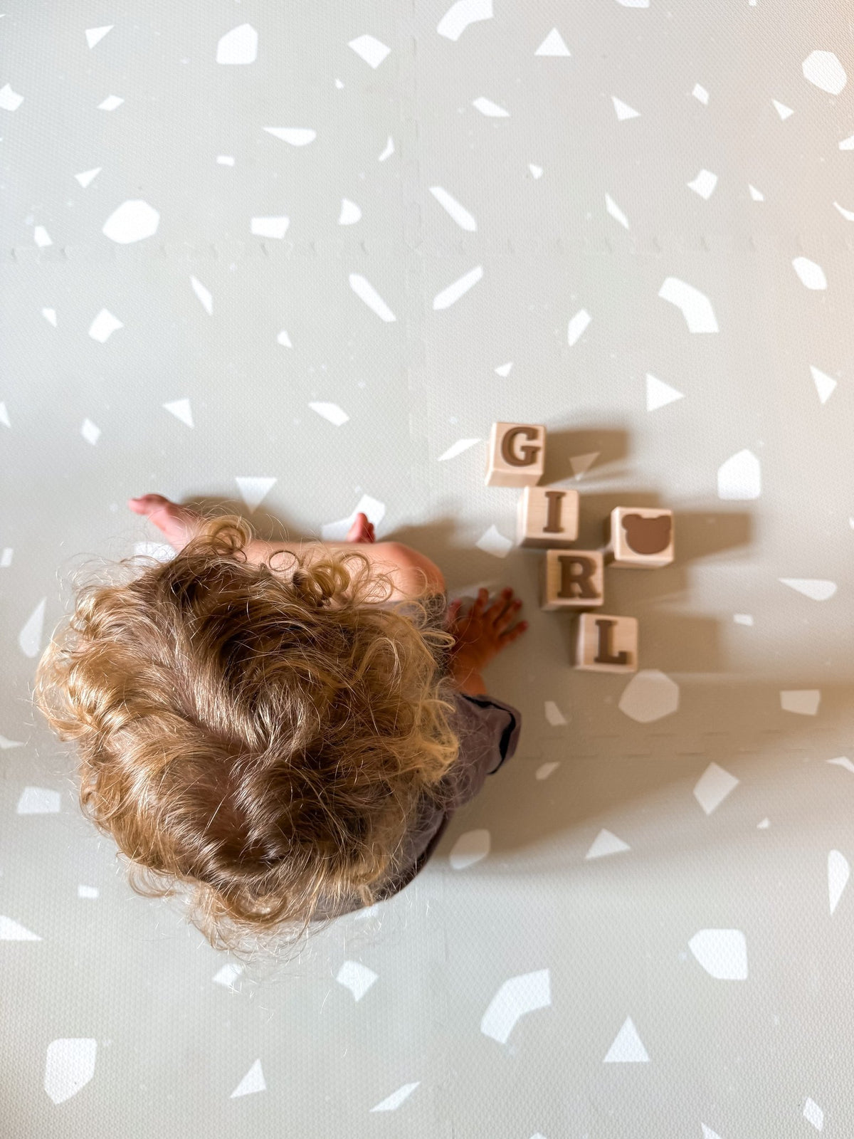 2. Top view of child sitting on OLYA KIDS Terrazzo Dove foam puzzle playmat with wooden blocks spelling 'GIRL'