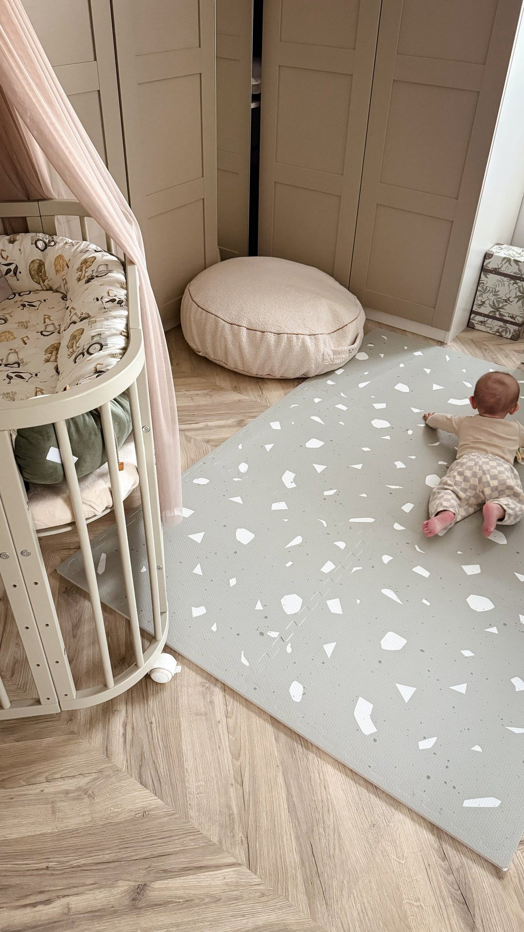 4. Baby on OLYA KIDS Terrazzo Sage playmat in nursery with beige crib and cushion, wooden floor.