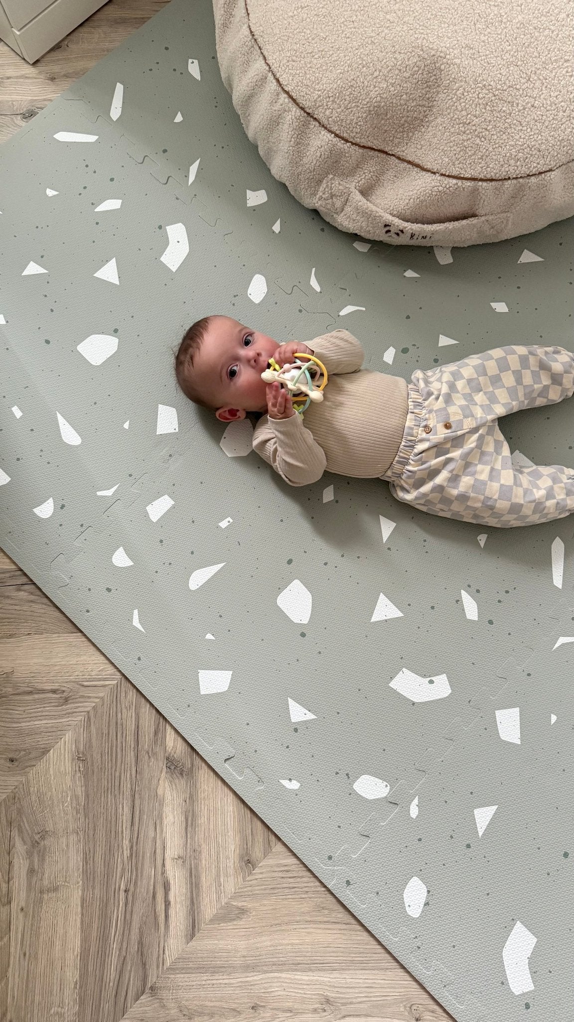 3. Baby on OLYA KIDS Terrazzo Sage playmat with beige cushion, wearing checkered pants, holding a toy.