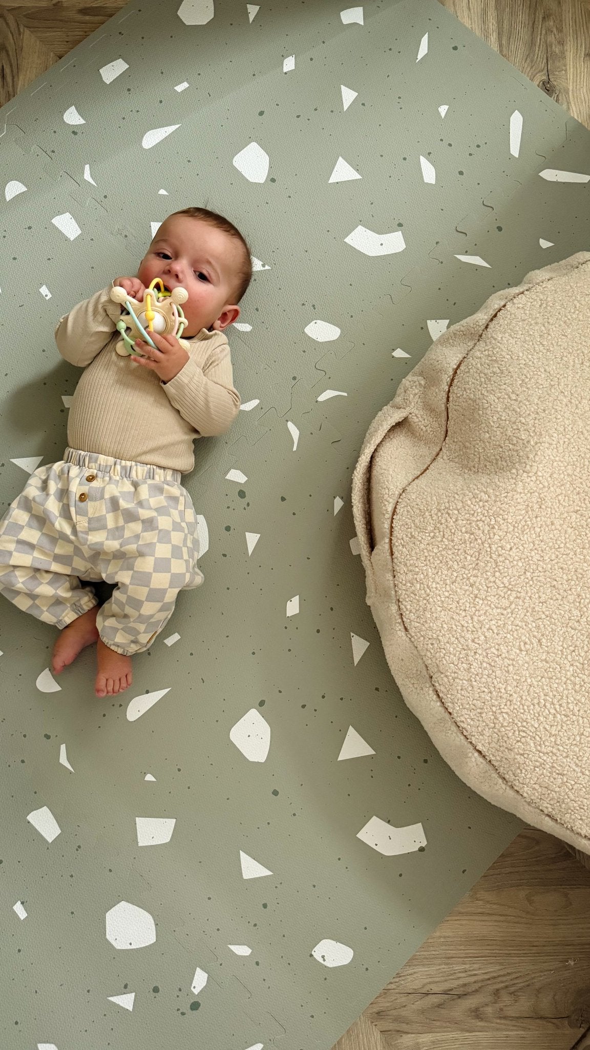 1. Baby lying on OLYA KIDS Terrazzo Sage foam playmat with beige and white checkered pants, holding a toy, next to a beige cushion.