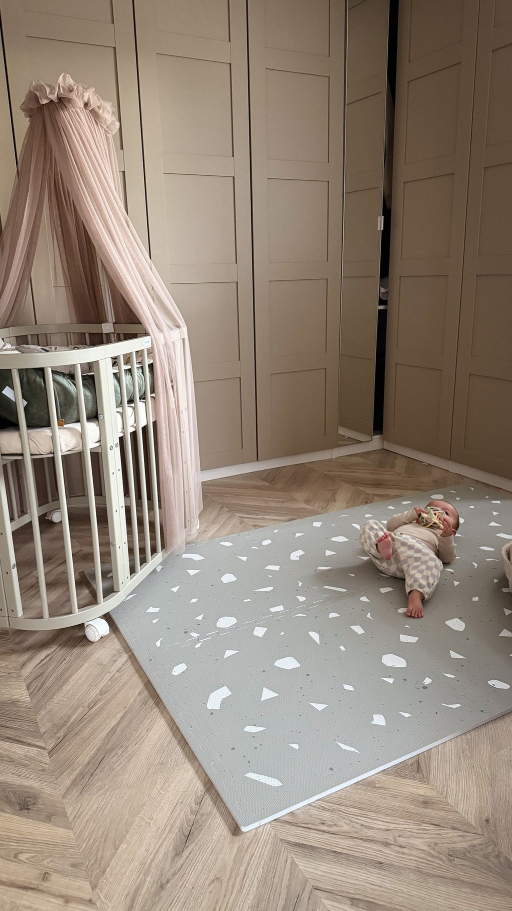 2. Baby on OLYA KIDS Terrazzo Sage playmat in nursery with beige crib and canopy, wooden floor.