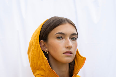 1. Woman wearing Lentsius Folia Mini Earrings in gold-tone with orange hooded jacket, light background