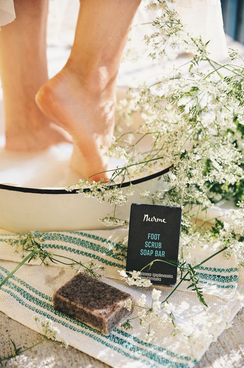 1. Woman soaking feet in basin with Nurme foot scrub soap bar on towel, surrounded by flowers, in a natural setting