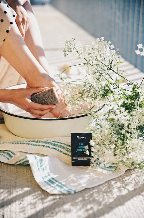 1. Woman using Nurme foot scrub soap bar on feet in basin, with flowers and towel, in a sunlit outdoor setting
