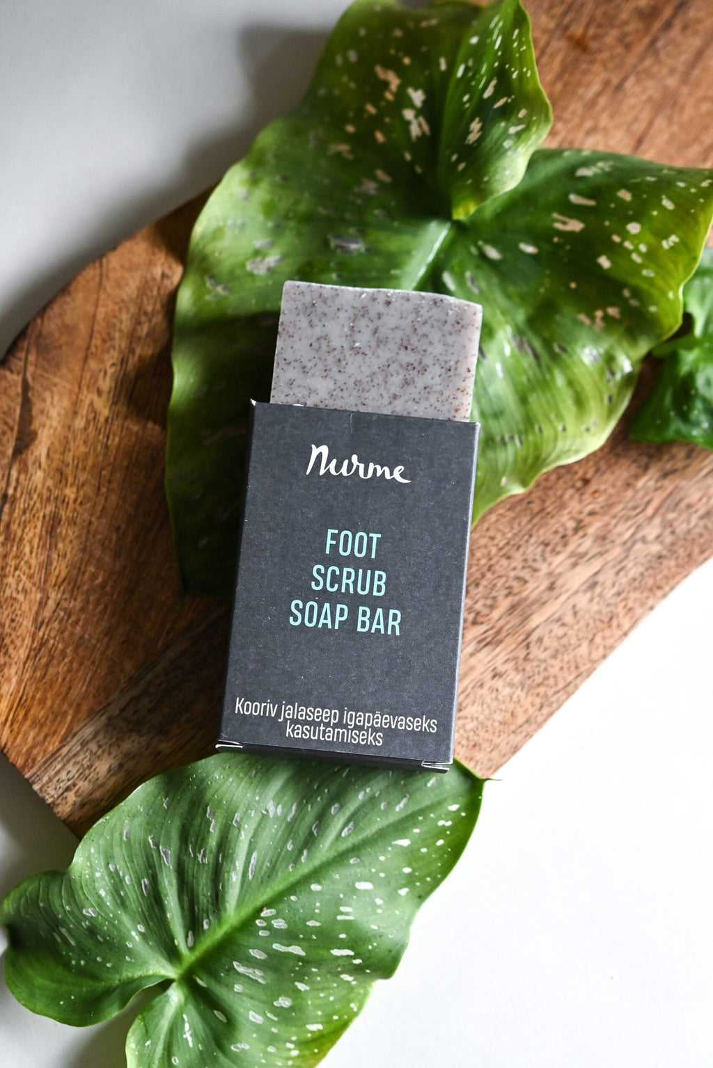 3. Close-up of Nurme foot scrub soap bar on wooden surface with green leaves, highlighting eco-friendly packaging
