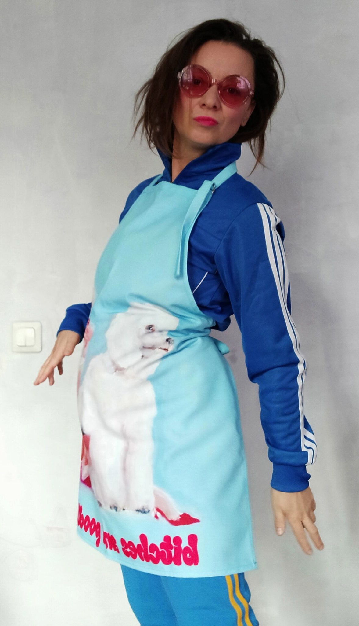 1. Woman wearing Katrin Valgemäe apron with poodle artwork, dressed in blue tracksuit and pink sunglasses