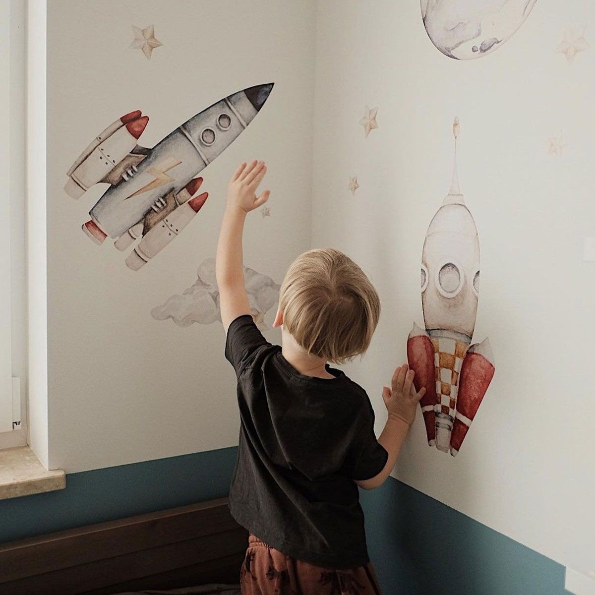 1. Child reaching towards watercolor rocket wall stickers in a space-themed room with stars and planets