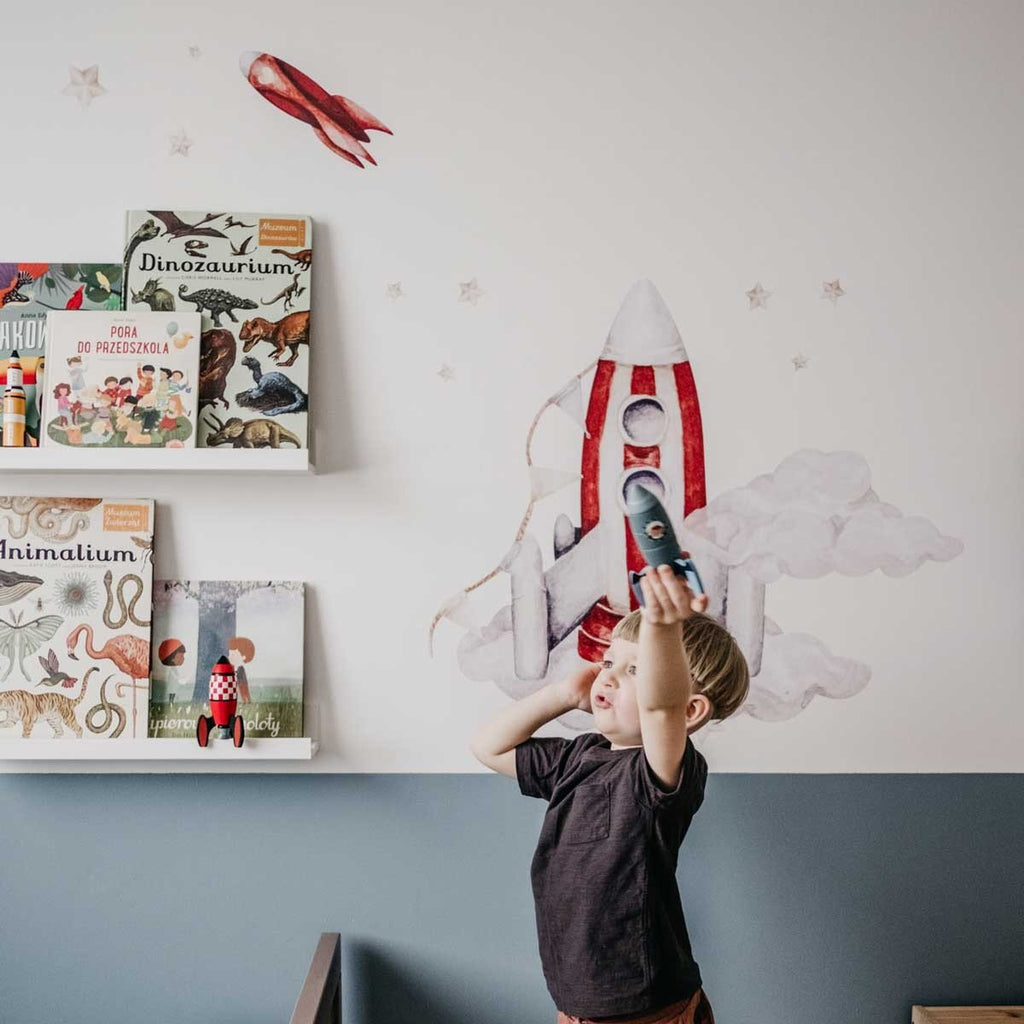 7. Child holding toy rocket in a room with watercolor rocket wall stickers and books on shelves