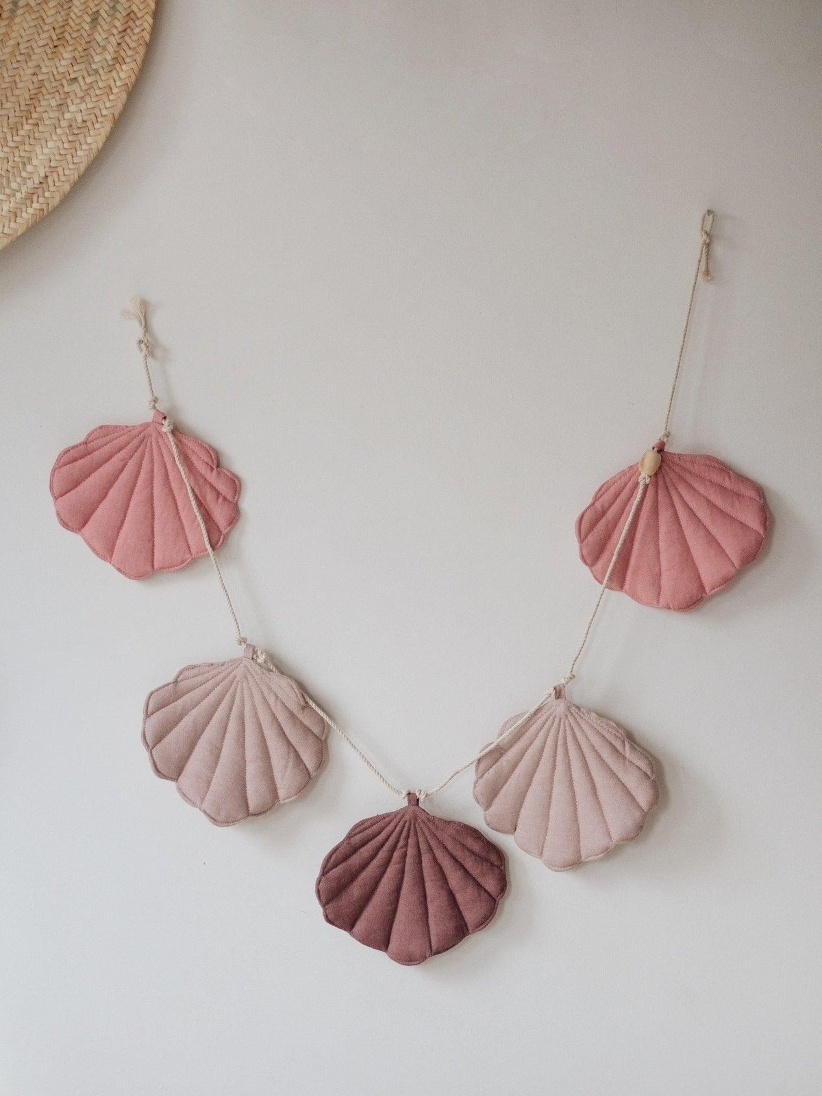 1. Powder pink shell garland with five linen pendants in various pink shades hanging on a wall with a straw hat and basket decor