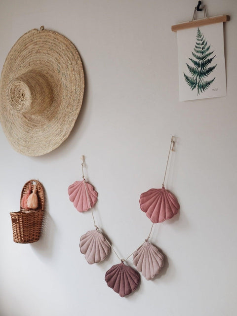 5. Powder pink shell garland with five linen pendants hanging on a wall with a straw hat and fern print decor
