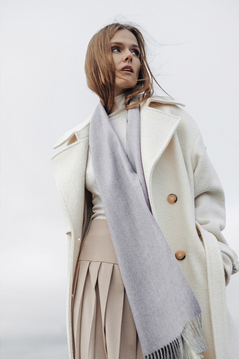 1. Woman wearing Alpaka natural baby alpaca wool scarf in light grey, styled with a white coat, outdoor setting