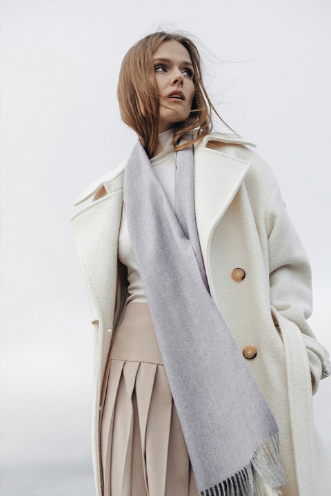 1. Woman wearing Alpaka natural baby alpaca wool scarf in light grey, styled with a white coat, outdoor setting