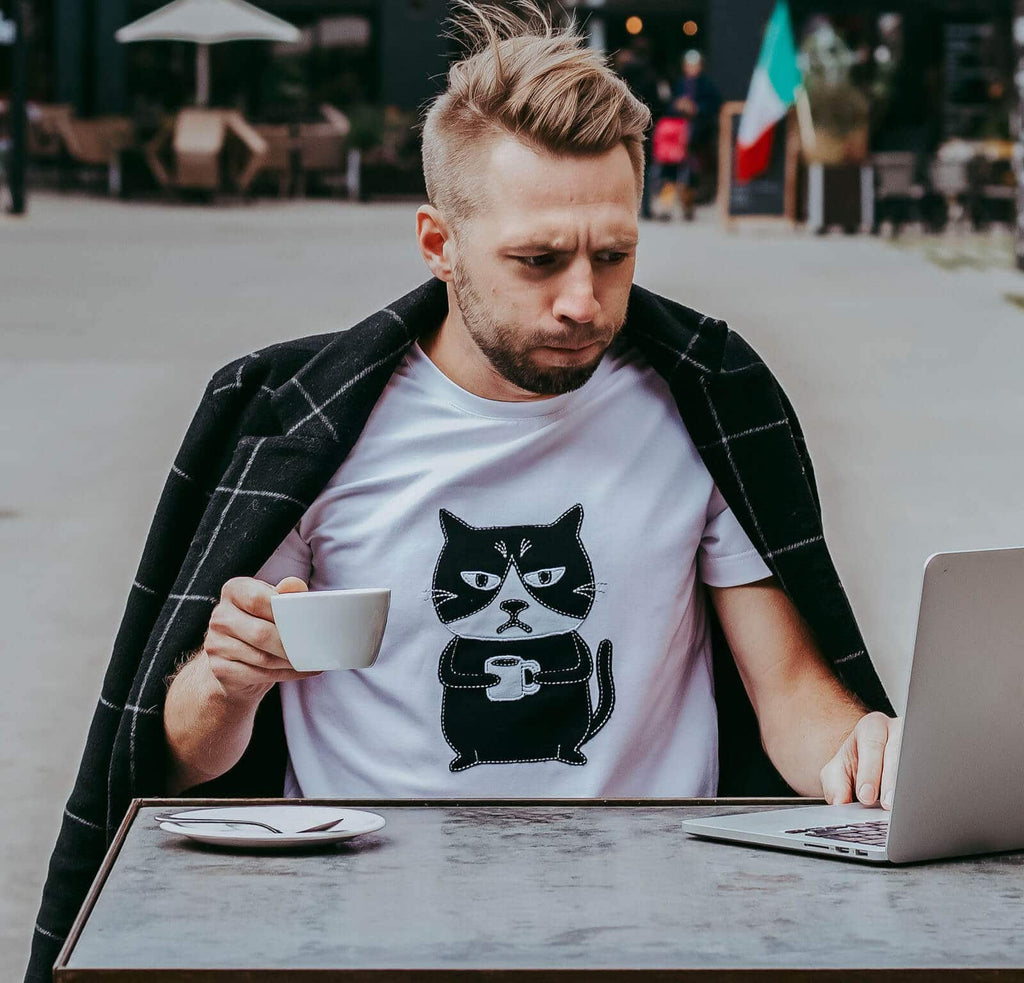 1. Men's Grumpy Cat T-shirt in white by HILP, worn by a male model enjoying coffee outdoors