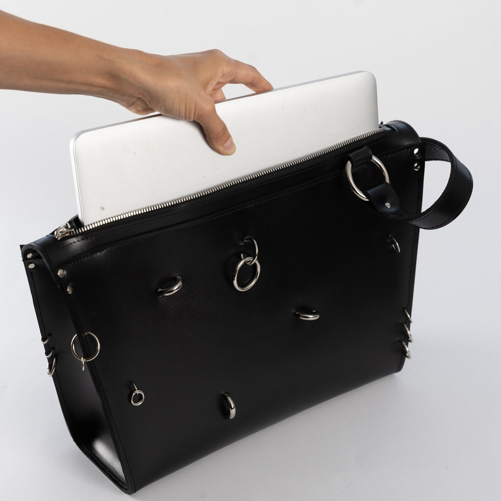 2. Hand placing laptop into black ARINA handbag with silver rings, demonstrating size and functionality