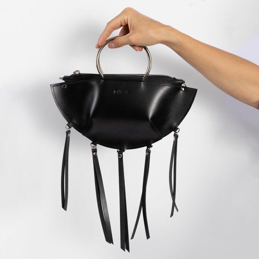 7. Hand holding black leather KAYA handbag with metal ring handles and tassels against a white background