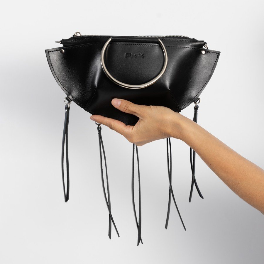 4. Hand holding black leather KAYA handbag with metal ring details and tassels against a white background