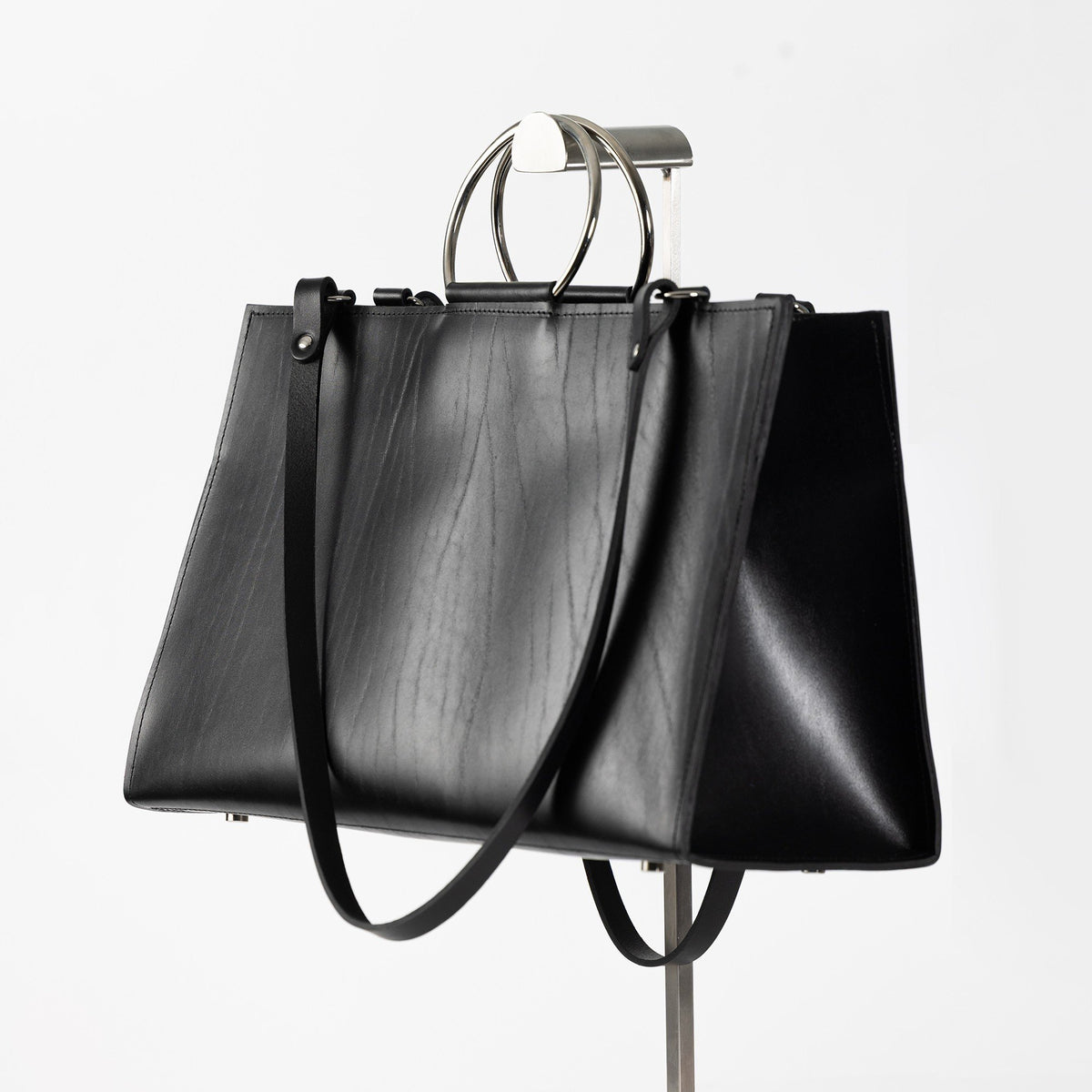 4. Black leather handbag with metal ring handles and shoulder straps, displayed on a stand in studio