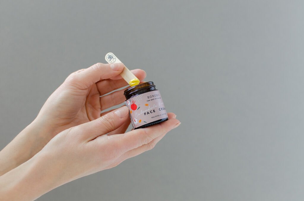 2. Hands holding BONOBO face cream jar with handcrafted juniper spatula, showcasing product use