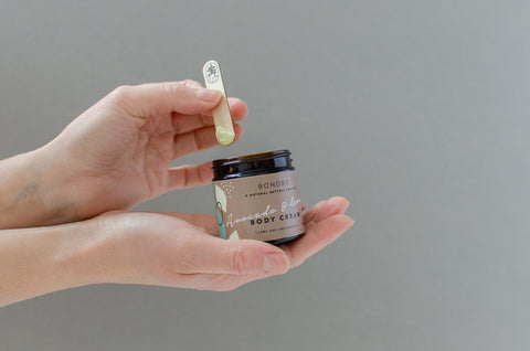 1. Hands holding BONOBO Avocado & Aloe Body Cream jar with wooden spatula, showcasing product texture