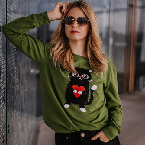 1. Woman wearing HILP Heartful Cat Sweatshirt in dark green, styled with sunglasses, outdoor setting