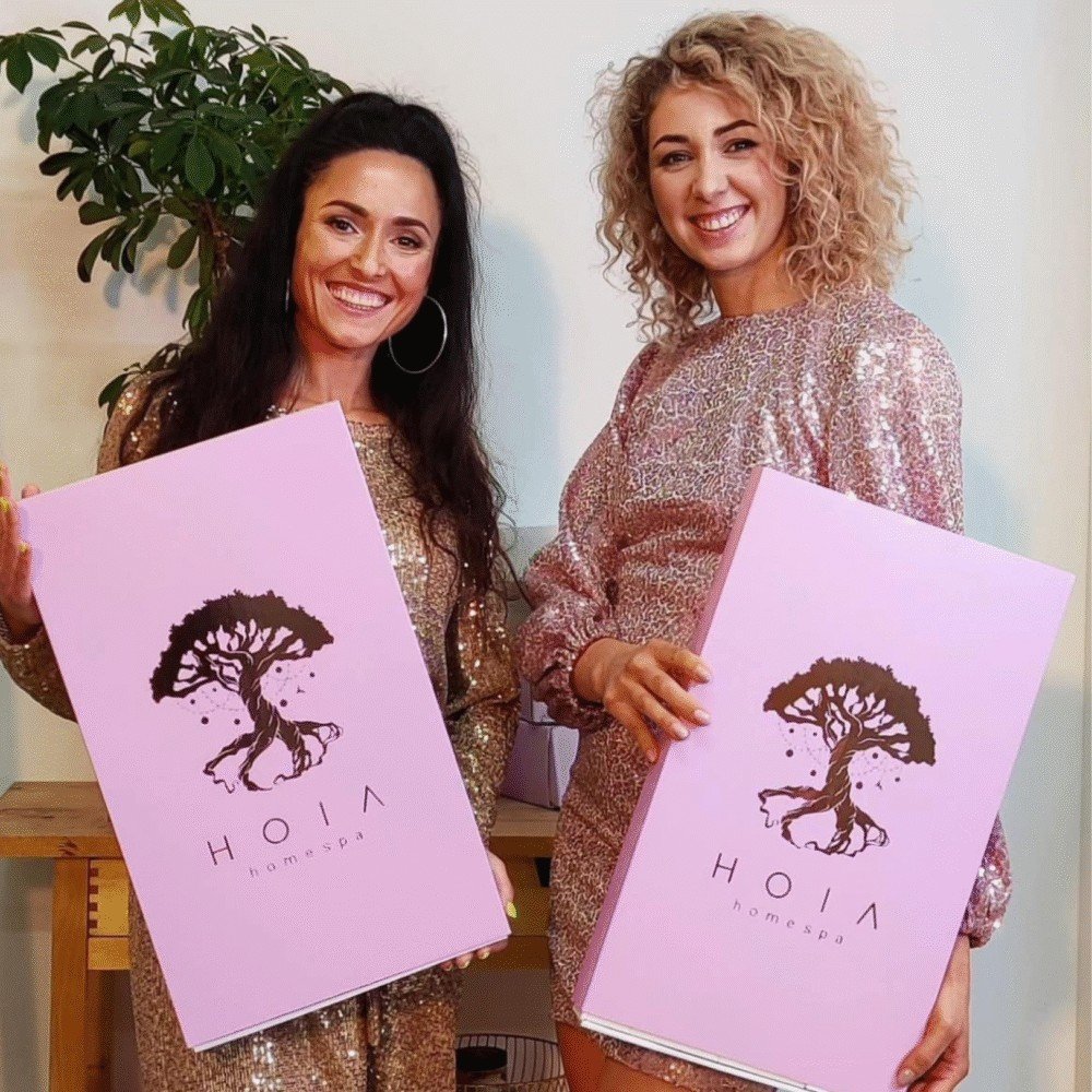 6. Two women holding HOIA Advent Calendar 2025 boxes, wearing sparkly dresses indoors