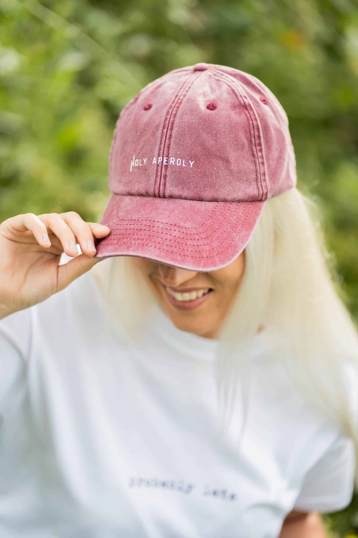 1. Woman wearing pastel pink Holy Aperoly cap by Black Giraffe Brand, smiling in outdoor setting