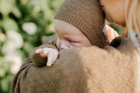 1. Baby wearing brown cable knit bonnet held by adult in outdoor setting