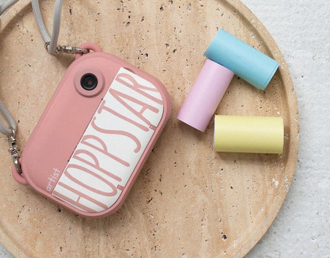 1. Hoppstar Artist camera in pink with three colorful thermal paper rolls on a wooden surface
