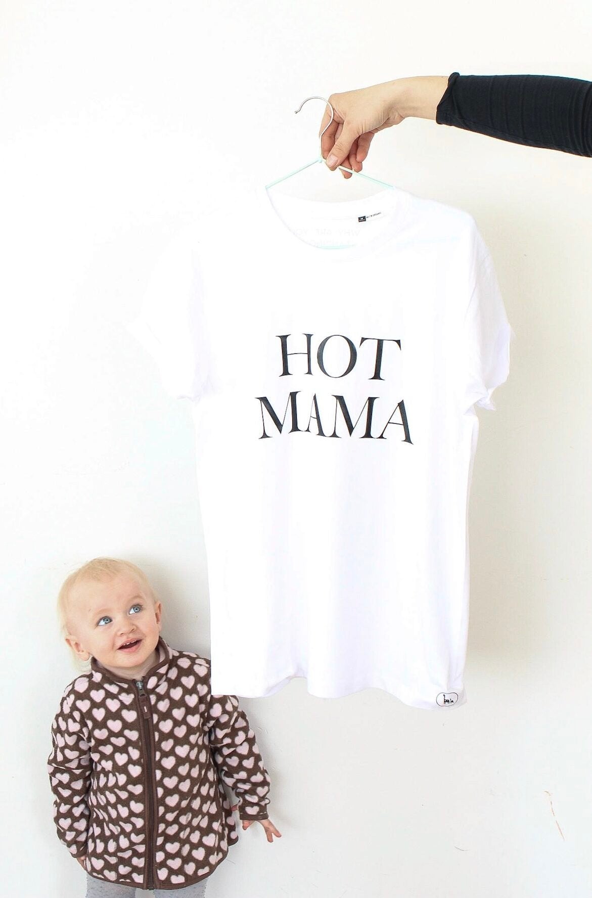 1. White Hot Mama T-shirt by Black Giraffe Brand, shown hanging with bold text, next to a child in a heart-patterned jacket