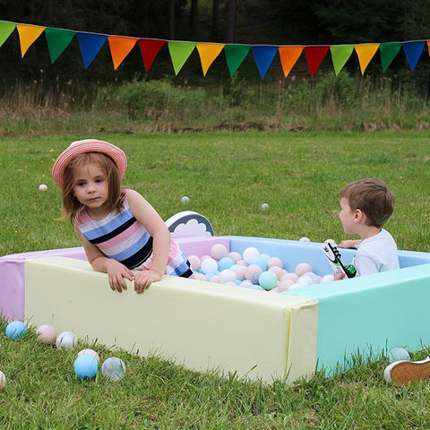 1. Two children playing in an outdoor ball pit filled with IGLU pastel balls, surrounded by colorful bunting and grass