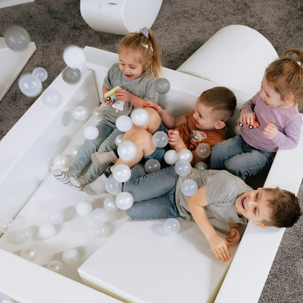 1. Three children playing in a white IGLU soft play ball pit with grey and white balls, indoors on a carpeted floor