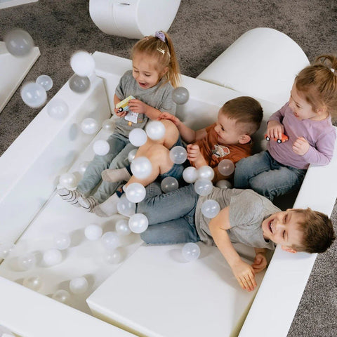 1. Three children playing in a white IGLU soft play ball pit with grey and white balls, indoors on a carpeted floor