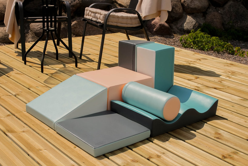 1. IGLU Discoverer 7-piece foam block set in light pastel colors arranged on a wooden deck outdoors with chairs and plants in the background