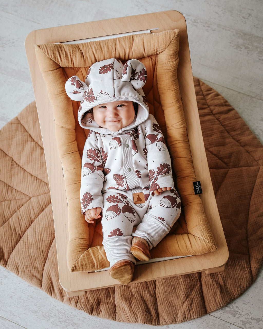 1. Baby wearing Zezuzulla eared jumpsuit with acorn print, lying on a cushioned mat