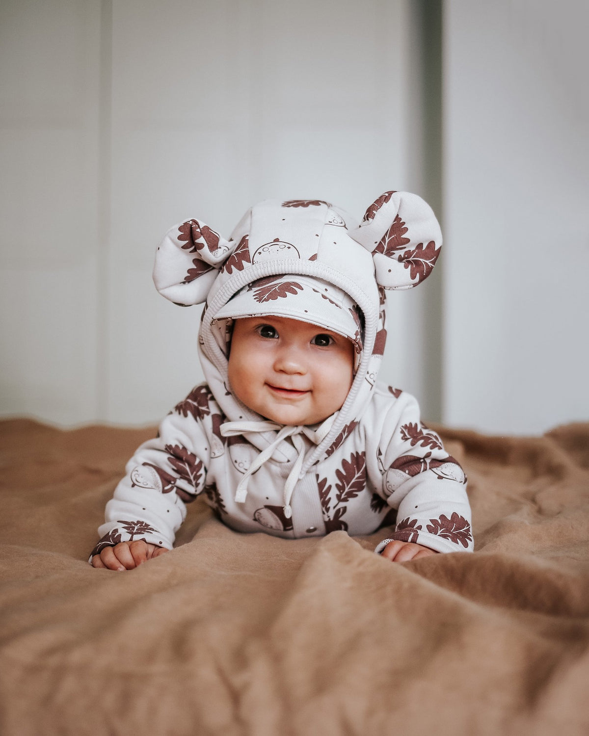 3. Baby in Zezuzulla eared jumpsuit with acorn print, smiling while lying on a bed