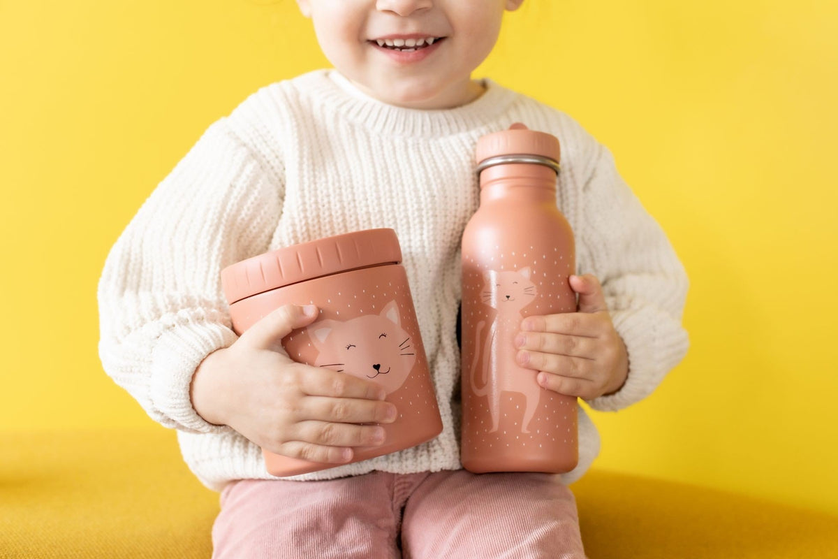 1. Child holding Trixie Baby insulated lunch pot and bottle with Mrs. Cat design, wearing white sweater and pink pants against yellow background