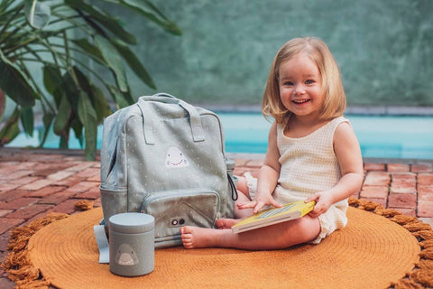 1. Young girl sitting on orange mat with Trixie Baby Mr. Shark lunch pot and matching backpack outdoors