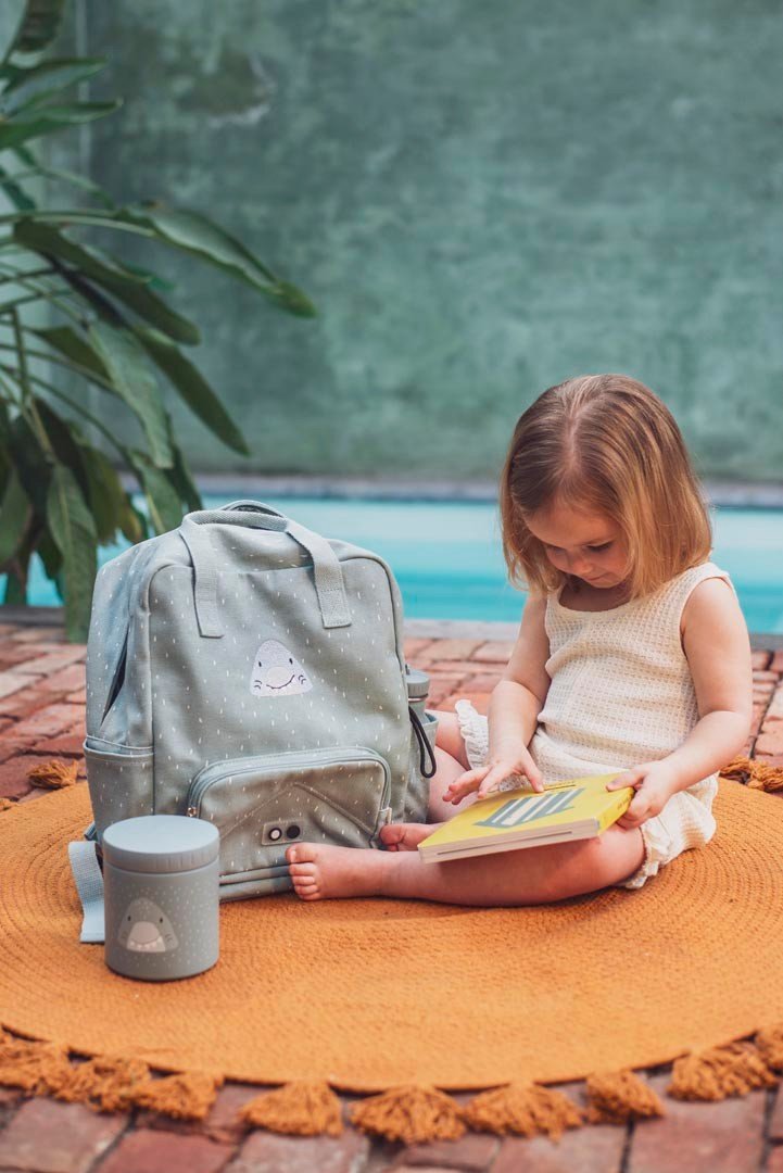 1. Young girl reading book next to Trixie Baby Mr. Shark lunch pot and backpack on orange mat outdoors