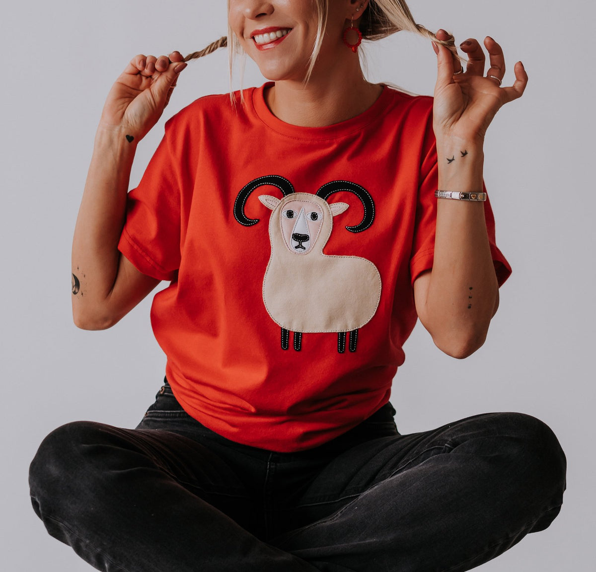 1. Women's red Aries T-shirt by HILP with ram graphic, worn by model sitting cross-legged