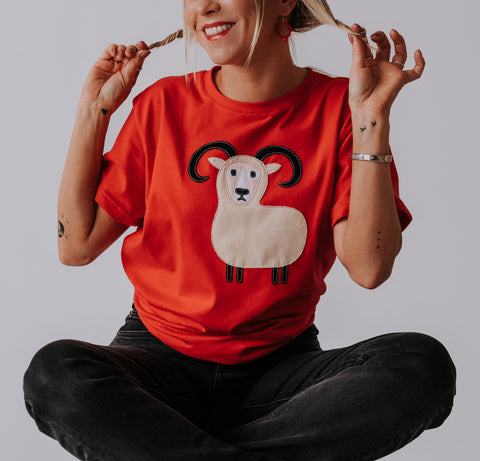 1. Women's red Aries T-shirt by HILP with ram graphic, worn by model sitting cross-legged