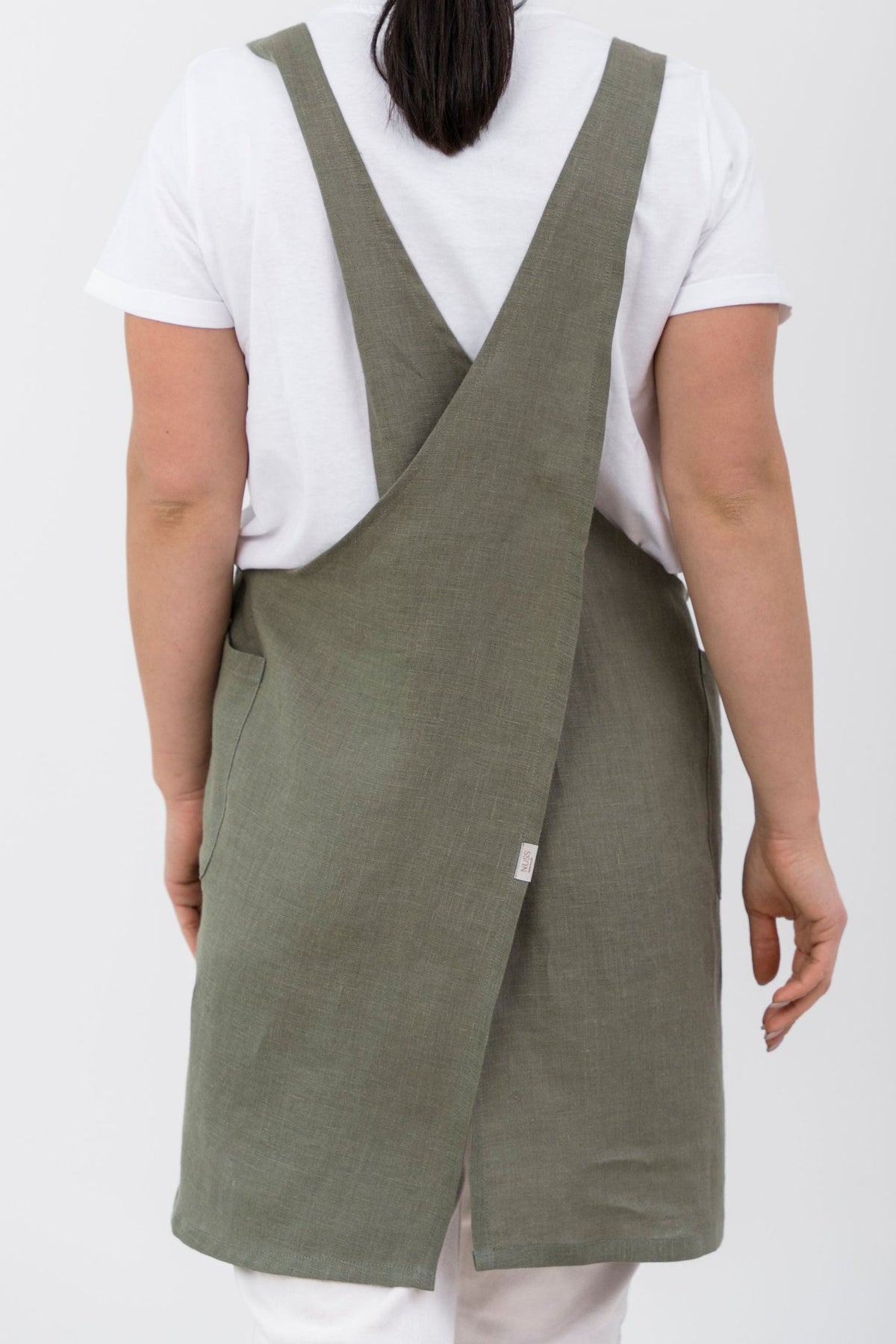 11. Woman wearing olive green Japanese-style linen apron with cross-back design, shown from back, over white shirt