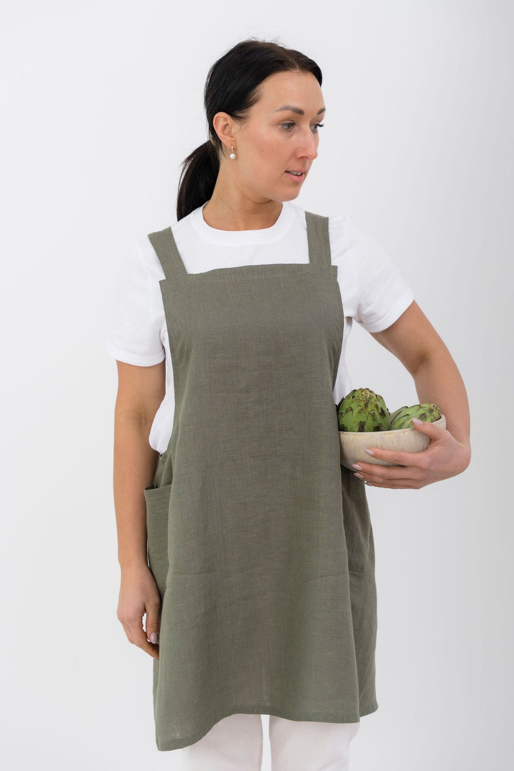 3. Woman in olive green Japanese-style linen apron holding artichokes, standing against white background